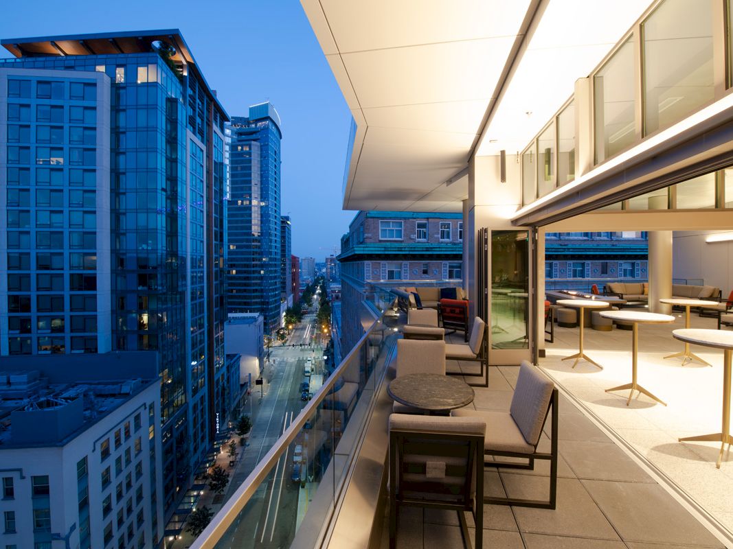 An elegant balcony with seating and tables overlooks a cityscape with high-rise buildings. The scene is captured during the evening.