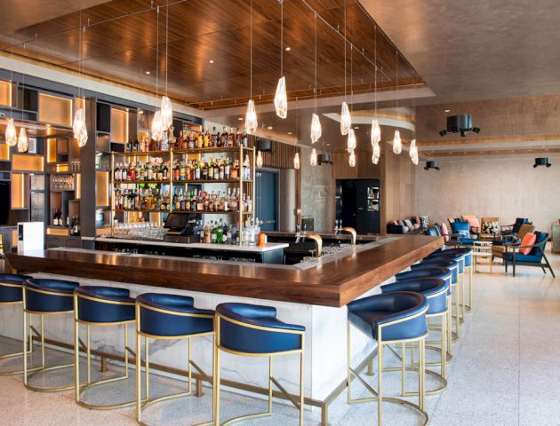 The image shows a modern bar with a large counter, high stools, and overhead pendant lights. There are shelves with various bottles behind the counter.