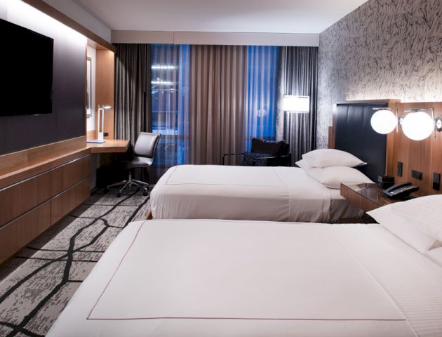 The image shows a modern hotel room with two beds, a TV on the wall, a working desk, lamps, and large windows with curtains.