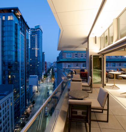 A rooftop balcony with seating overlooks a city skyline at dusk. Buildings are lit, and the open area has tables and chairs set up.