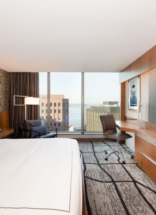 The image shows a modern hotel room with a large bed, a desk with a chair, a flat-screen TV, and a window with a view of buildings and the water.