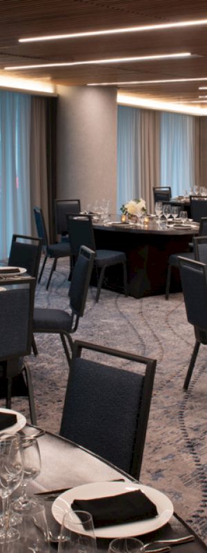 A banquet room with round tables set for an event, featuring black chairs, neatly arranged place settings, and soft lighting.