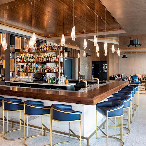 The image shows an upscale bar with a wooden counter, blue bar stools, and pendant lighting, featuring a well-stocked bar and cozy seating areas.