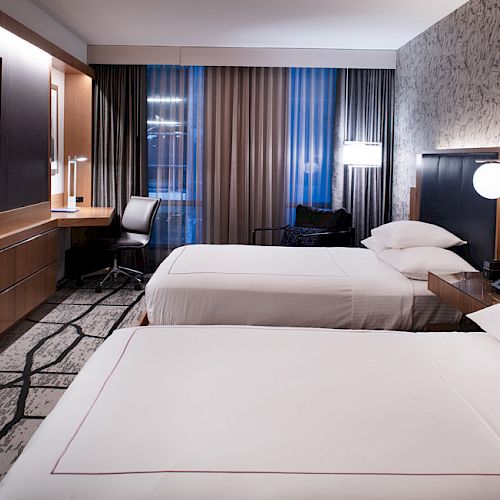 The image shows a modern hotel room with two beds, a TV on the wall, a desk with a chair, and stylish decor.