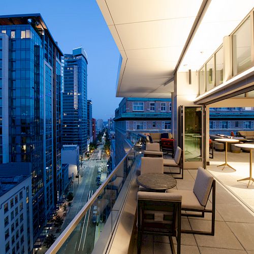 An evening cityscape view from a modern apartment balcony, featuring outdoor seating and adjacent indoor area with tables and lighting.