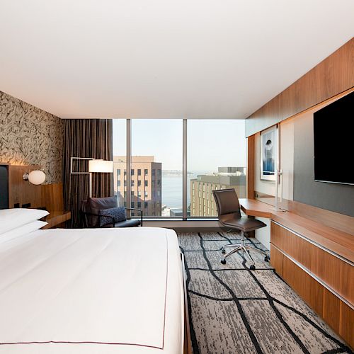 A modern hotel room features a large bed, wall-mounted TV, desk with a chair, and a window view of a cityscape with buildings and water.