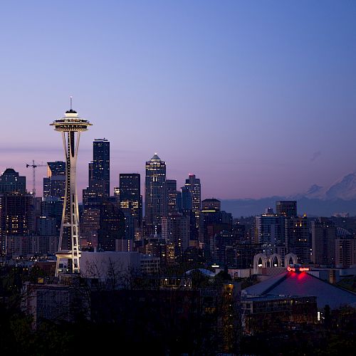 The image shows a nighttime skyline of Seattle, featuring the Space Needle prominently with Mount Rainier visible in the background.