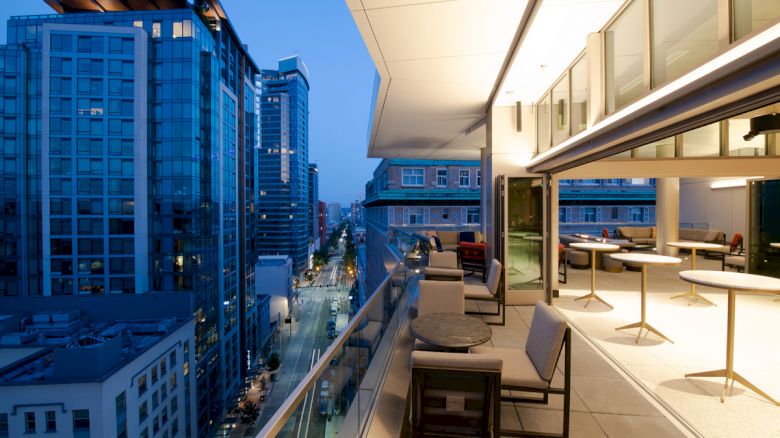 The image shows a modern rooftop terrace at dusk, with seating areas and city skyscrapers in the background. The view includes a busy street below.