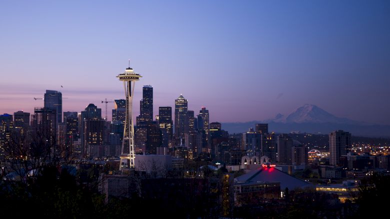 The image depicts the Seattle skyline at dusk, featuring the Space Needle and Mount Rainier in the background, with city lights beginning to illuminate.