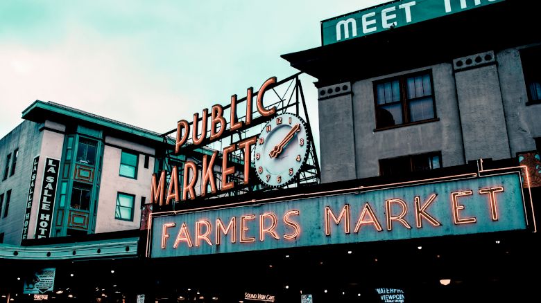 This image shows the entrance of a Public Market with neon signs that read "Farmers Market" and a clock, surrounded by buildings.