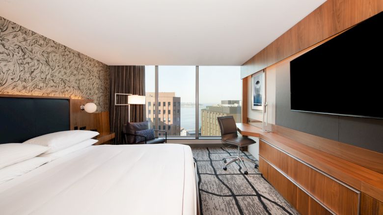 A modern hotel room with a large bed, a wall-mounted TV, a desk with a chair, and a view of buildings through the window.