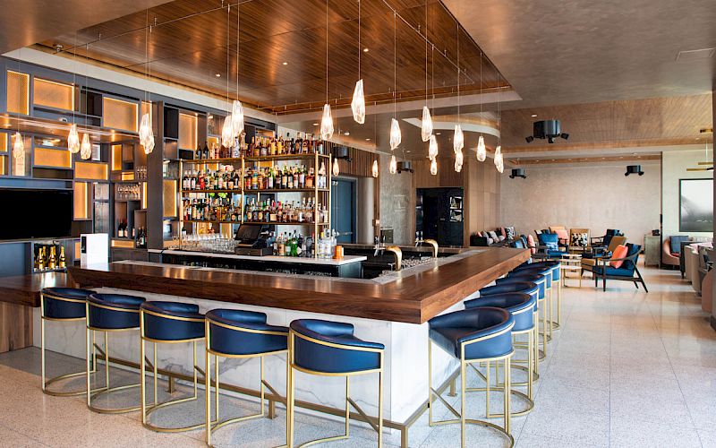 The image shows a modern bar with a countertop, bar stools, hanging lights, and a wide selection of bottles behind the counter, all in a stylish setting.