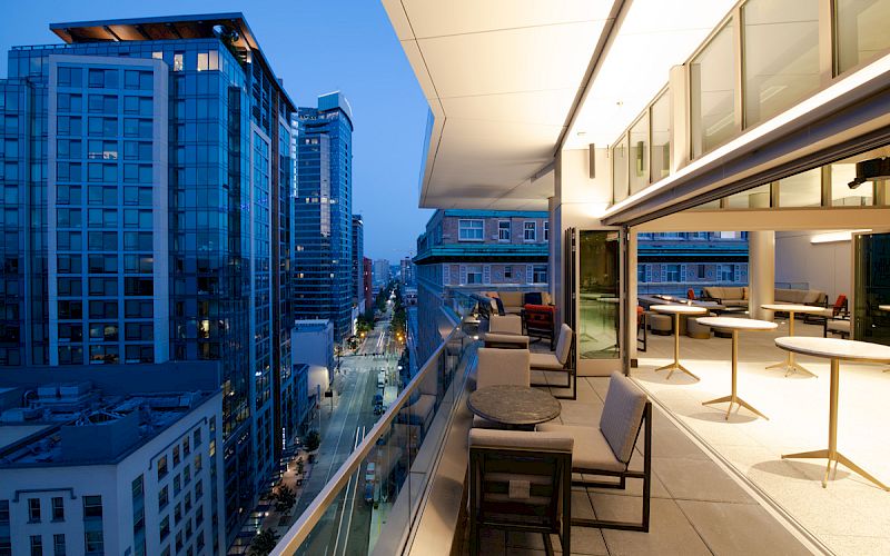 A modern balcony with seating and tables, overlooking a cityscape with tall buildings and illuminated streets at twilight.