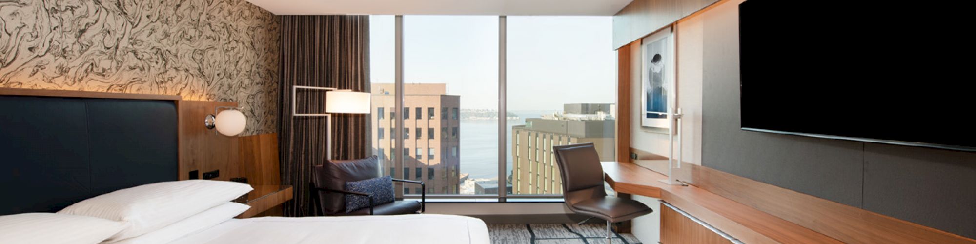 A modern hotel room with a large bed, a sleek desk and chair, a flat-screen TV, and a city view through a window, featuring contemporary decor.