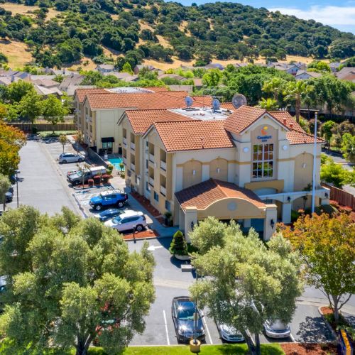 The image shows a Comfort Inn hotel surrounded by trees and hills, with a parking lot filled with cars and a clear blue sky above.