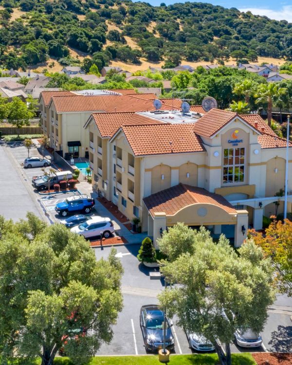 The image shows a Comfort Inn hotel surrounded by trees and hills, with a parking lot filled with cars and a clear blue sky above.