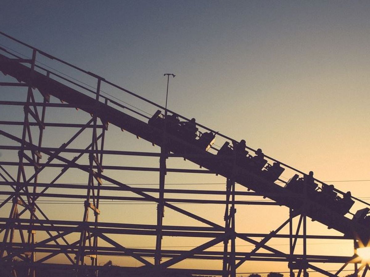 The image depicts a roller coaster with silhouetted riders ascending a steep incline against a backdrop of a sunset or sunrise.