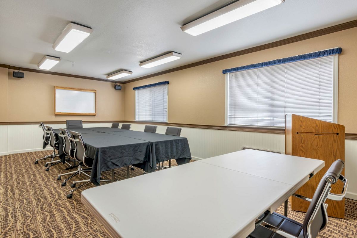 A small conference or meeting room with a long table covered with a black cloth, surrounded by chairs, and two windows with blinds.