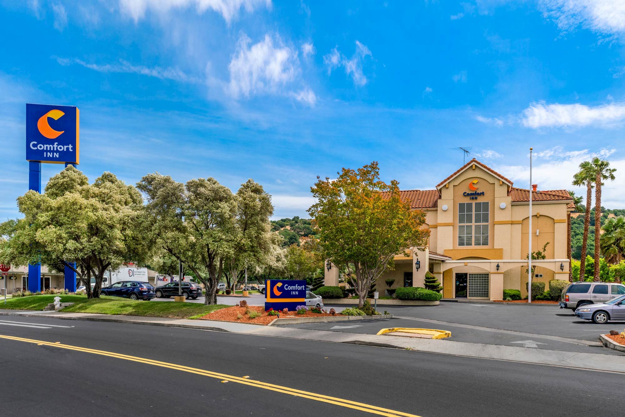 The image shows a Comfort Inn hotel with a sign, trees, a parking lot, and a clear blue sky, located near a road intersection.