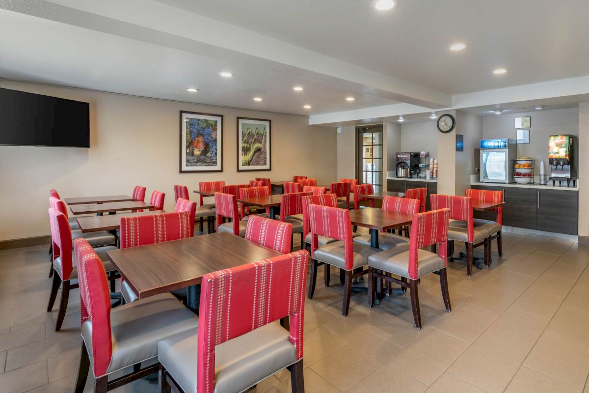 The image shows a clean, modern dining area with wooden tables, red and gray chairs, wall art, and vending machines by the far wall under soft lighting.